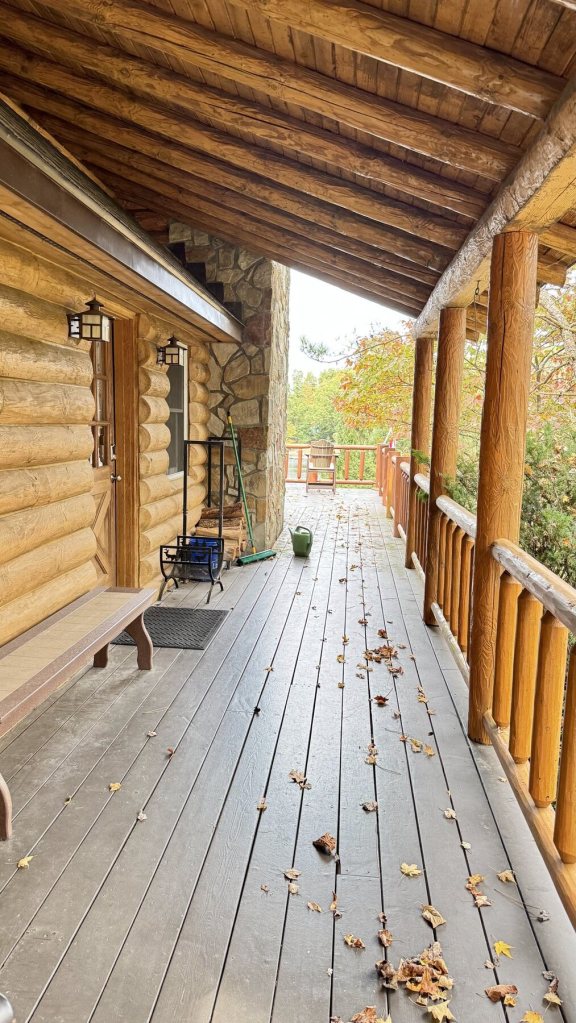 Long covered porch with log posts, a bench by the door and fallen leaves on the deck boards
