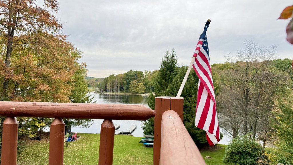 View from a log deck railing with an American flag, looking down at the lake through fall trees