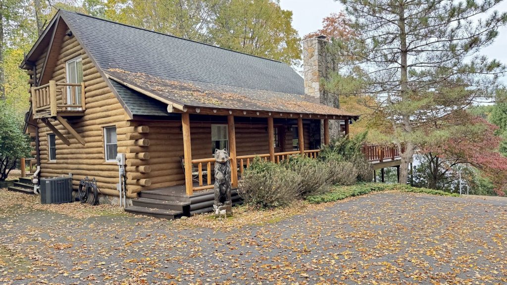 Log cabin with a stone chimney, covered front porch and a carved wooden bear by the steps, surrounded by fall leaves and pine trees