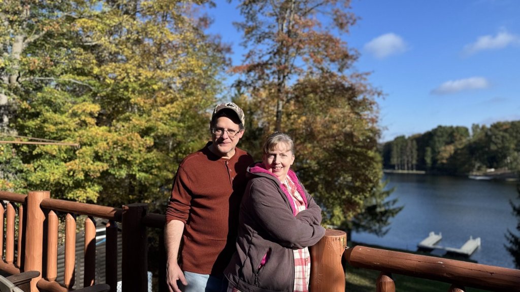Corilee and Grant standing on the log cabin deck with the lake and blue sky behind them
