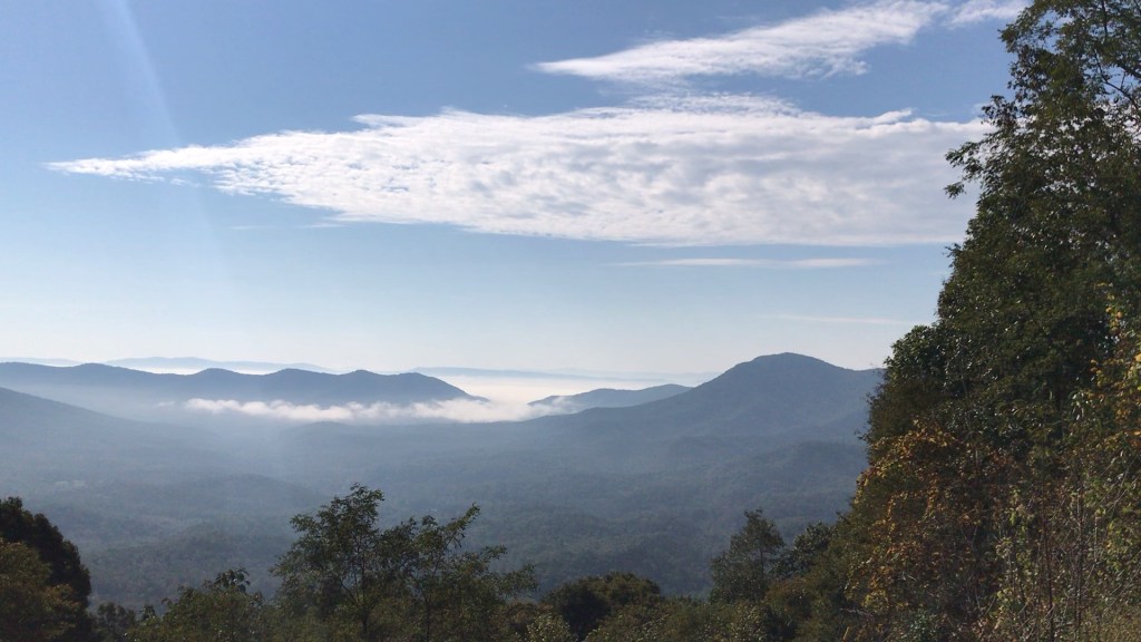 Layered blue mountain ridges with fog settled in the valleys below a Blue Ridge Parkway overlook, under a blue sky with a long streak of white cloud