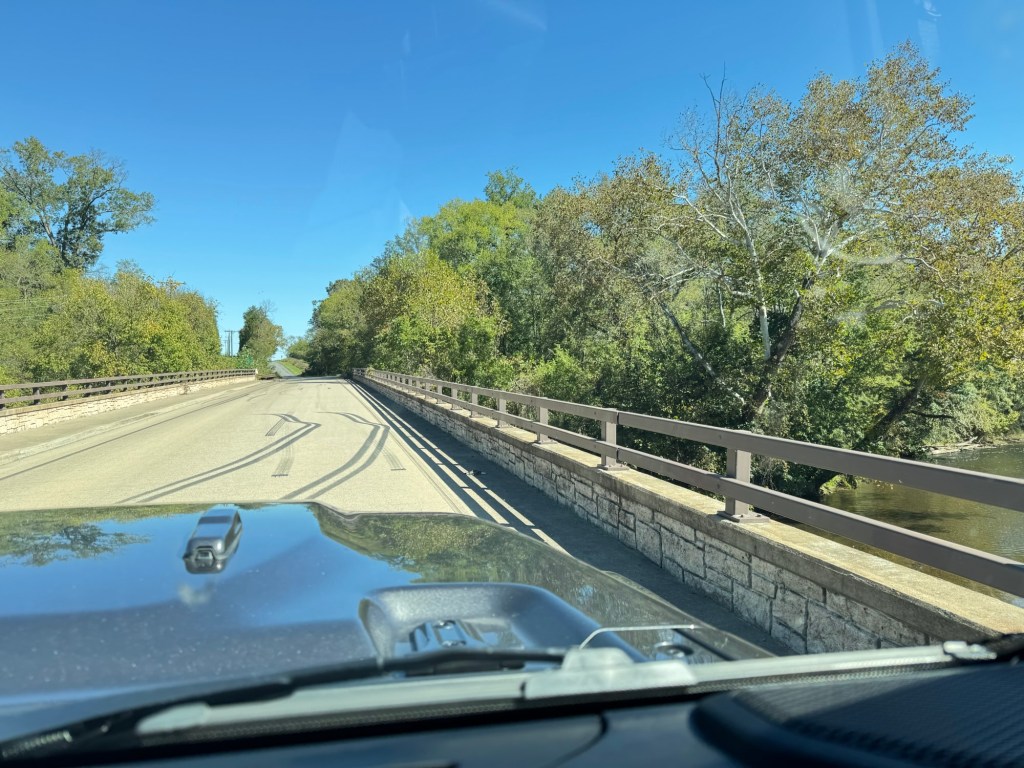 View over the hood while driving onto a bridge with a stone wall and guardrail, trees on both sides