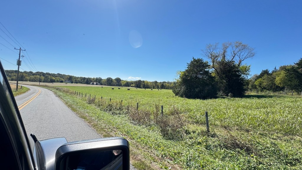Two-lane country road curving past a fenced green pasture under a clear blue sky, seen from the car window